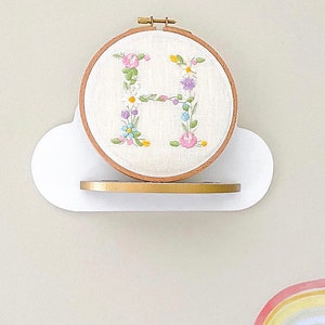 May include: A white cloud-shaped shelf with a gold trim holds a wooden embroidery hoop with a white fabric background. The fabric is embroidered with a floral design that spells out the letter "H".