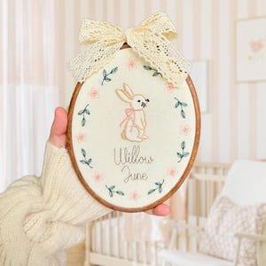May include: An oval embroidery hoop with a stitched bunny wearing a pink bow, encircled by floral and leaf details. The text "Willow June" is embroidered beneath the bunny. A cream lace bow is attached at the top. The hoop is displayed in a nursery.