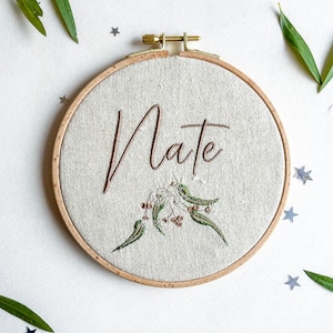 May include: A round embroidery hoop with a beige linen fabric background. The name "Nate" is embroidered in brown cursive script with a small floral design below it.