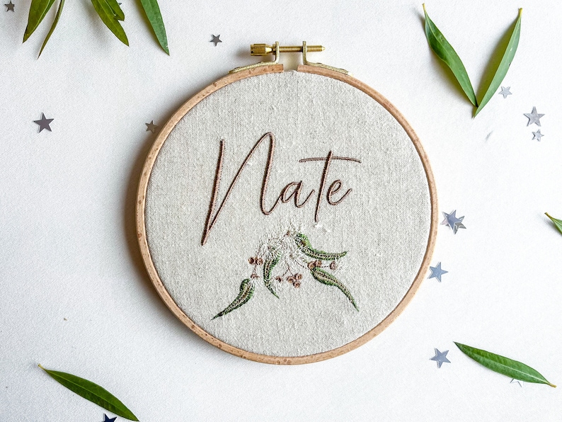 May include: A round embroidery hoop with a beige linen fabric background. The name "Nate" is embroidered in brown cursive script with a small floral design below it.