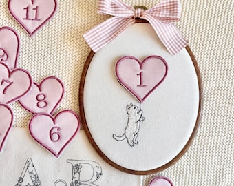 Könnte beinhalten: Ein ovaler Holzstickrahmen mit weißem Stoff, auf dem eine weiße Katze gestickt ist, die ein rosa Herz mit der Zahl 1 hält. Oben ist eine rosa karierte Schleife befestigt. Mehrere lose rosa Herzaufnäher mit den Zahlen 2, 6, 7, 8, 9 und 11 liegen daneben, sowie ein Stoffstück mit dem Text A&B ARCHER AND BOO.