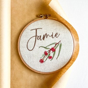 May include: A round embroidery hoop with a beige fabric background. The name "Jamie" is embroidered in brown cursive script. A sprig of green leaves with red flowers is embroidered below the name.