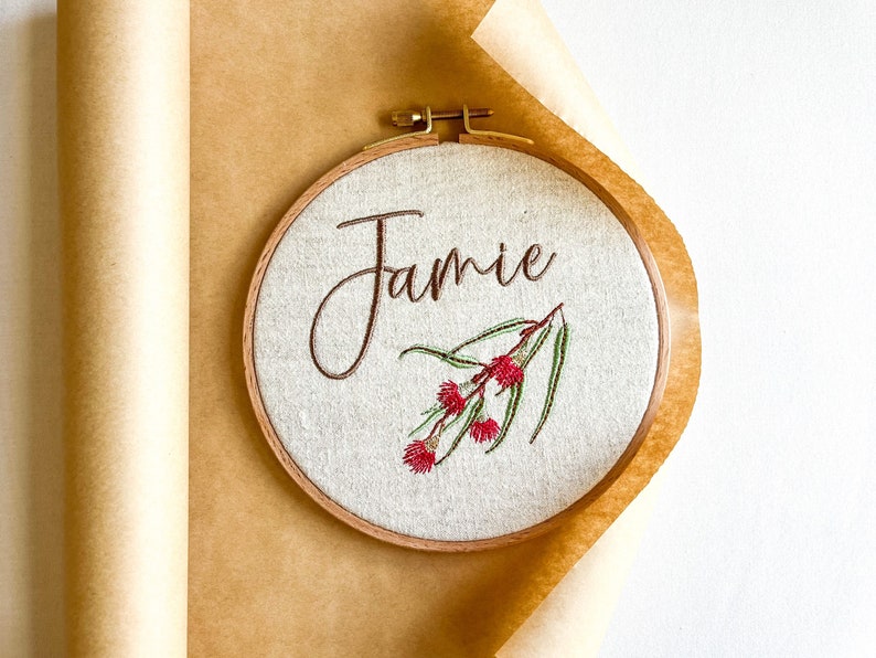 May include: A round embroidery hoop with a beige fabric background. The name "Jamie" is embroidered in brown cursive script. A sprig of green leaves with red flowers is embroidered below the name.