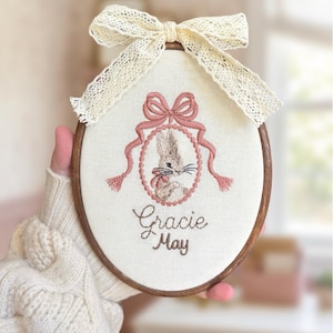 May include: An oval wooden embroidery hoop containing off-white fabric with a stitched illustration of a rabbit inside a pink oval frame topped with a pink bow. Below the frame, the name Gracie May is embroidered in brown. A cream lace bow sits on top.