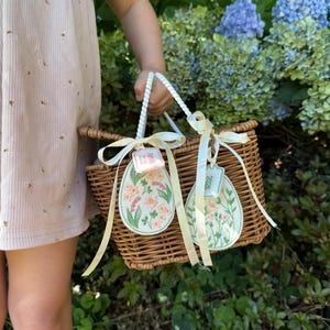 May include: A wicker basket with white ribbon handles and two embroidered egg-shaped tags. The tags are decorated with floral designs and the text "2023".
