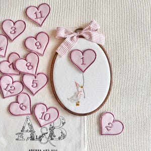 May include: An embroidered nursery hoop featuring a goose holding a heart-shaped balloon with the number 1. Pink heart-shaped patches with numbers 2-12 are scattered around. A fabric bag with the text "ARCHER AND BOO" is also present.