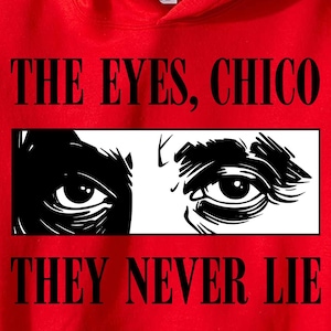 May include: Red hooded sweatshirt with the text "THE EYES, CHICO THEY NEVER LIE" in black. The design includes a black and white illustration of two eyes within a white rectangle.