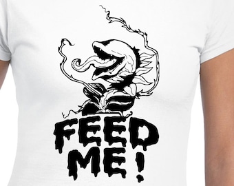 Puede incluir: Una camiseta blanca con una ilustración en blanco y negro de un monstruo vegetal con el texto "FEED ME!" impreso en ella.