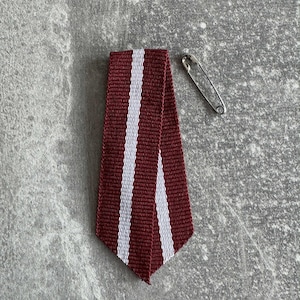 Op de afbeelding: Een bordeauxrood en wit gestreept lint met een veiligheidsspeld. Het lint is in een driehoekige vorm gevouwen. De veiligheidsspeld is zilverkleurig en bevindt zich bovenaan het lint.