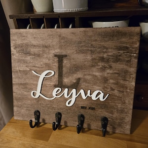 May include: A wooden key holder with the name "Leyna" and the date "EST. 2025" engraved on it. The key holder has four black metal hooks.