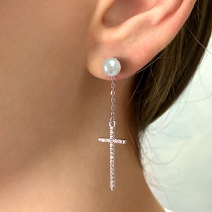 Puede incluir: Pendientes de plata con una perla en la parte superior y una cruz con piedras transparentes en la parte inferior.