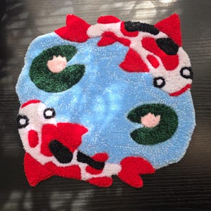 May include: A handmade rug featuring a pond scene with two koi fish and lily pads. The fish are red, white, and black, set against a light blue background. The lily pads are green with pink accents. The rug is a unique, decorative item.