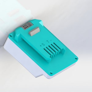 Può includere: Un pacco batteria turchese con un'etichetta bianca che dice '+ S.T.' e un piccolo interruttore nero nella parte superiore.
