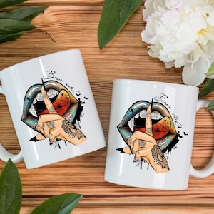 May include: Two white ceramic mugs with a Halloween-themed design. The design features a pair of blue and orange lips with fangs, a finger to the lips gesture, and the text "Basic witch". The mugs are on a wooden surface.