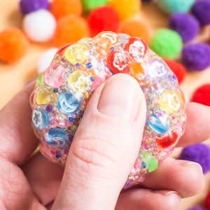 Könnte beinhalten: Ein farbenfroher, strukturierter Sensorikball mit eingebetteten fruchtförmigen Anhängern und kleinen Perlen. Der Ball wird in einer Hand gehalten, mit einem verschwommenen Hintergrund aus verschiedenfarbigen Pompons. Die Oberfläche des Balls ist eine Mischung aus Rosa, Gelb, Blau und Rot.
