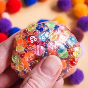 Op de afbeelding: Een kleurrijke stressbal gevuld met kleine, heldere kralen en verschillende gekleurde vormen. De bal wordt in een hand gehouden, met een vage achtergrond van kleurrijke pompons. De stressbal heeft een verscheidenheid aan kleuren, waaronder rood, oranje, geel, groen en blauw.