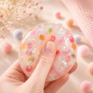 Könnte beinhalten: Ein kreisförmiges, durchscheinendes Fidget-Spielzeug aus Harz mit einer glatten, glänzenden Oberfläche. Das Objekt ist mit kleinen, bunten eingebetteten Perlen in Rosa-, Orange- und Gelbtönen sowie irisierenden Glitzerflocken gefüllt.