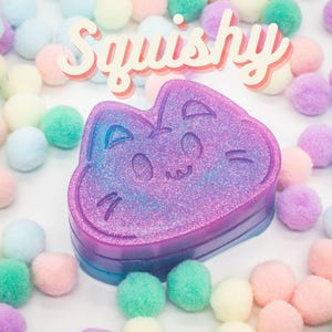 Può includere: Un giocattolo squishy a forma di gatto, di colore viola e blu con glitter e un viso carino. Il giocattolo è circondato da pompon colorati.