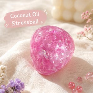 Könnte beinhalten: Ein durchscheinender, leuchtend pinker Stressball mit einer texturierten Oberfläche, die kleine kreisförmige Vertiefungen und eingebetteten Glitzer aufweist. Eine pinke Grafik oben links zeigt den weißen Text Coconut Oil Stressball.