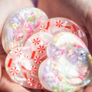 Può includere: Opere d'arte in resina a forma di cuore con caramelle colorate, zuccherini e personaggi dei cartoni animati incorporati. La resina trasparente permette di vedere il contenuto, con uno sfondo glitterato. I pezzi sono tenuti in una mano.
