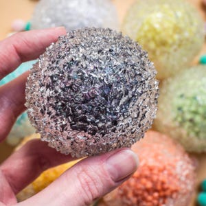Op de afbeelding: Een zilveren bol met een getextureerd oppervlak wordt in een hand gehouden, met andere kleurrijke bollen op de achtergrond. De bol heeft een ruwe, metalen uitstraling. De bollen op de achtergrond zijn in tinten geel, groen en oranje.