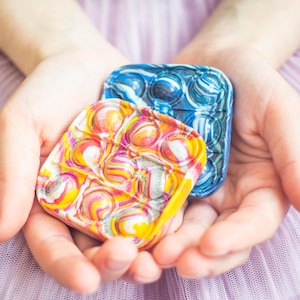 Op de afbeelding: Twee vierkante sensorische speeltjes van siliconen met een gemarmerde afwerking. De ene heeft een levendige werveling van geel, roze en wit met zilveren glitters, de andere een blauw-wit marmerpatroon, beide met cirkelvormige bubbels.