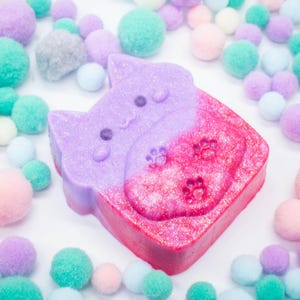 Könnte beinhalten: Eine zweifarbige katzenförmige Seife mit glitzerndem Finish. Die obere Hälfte ist lavendelfarben und die untere Hälfte ist ein leuchtendes Pink. Pfotenabdrücke sind auf dem rosafarbenen Abschnitt eingeprägt. Pastellfarbene Pompons umgeben die Seife.