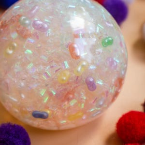 Könnte beinhalten: Ein klarer, runder sensorischer Ball, gefüllt mit bunten, geleeartigen Perlen und schillerndem Glitzer. Der Ball hat ein weiches, durchscheinendes Aussehen mit einer Vielzahl von Pastellfarben, darunter Rosa, Gelb und Lila. Andere bunte Pompons sind im Hintergrund zu sehen.