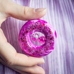 Op de afbeelding: Een cirkelvormige donut-bedel met een levendige magenta en roze gemarmerde basis. Het bovenoppervlak is zwaar ingelegd met roze en zilveren glitterdeeltjes van verschillende groottes. Een kleine zilveren speld houdt twee doorschijnende roze kralen vast.
