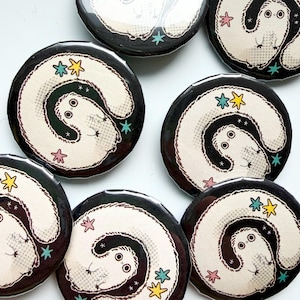 May include: A collection of circular pin-back badges featuring a black background with a central illustration of a white, long-bodied creature with large eyes. The design includes small, colourful star-shaped accents in yellow, teal, and pink.