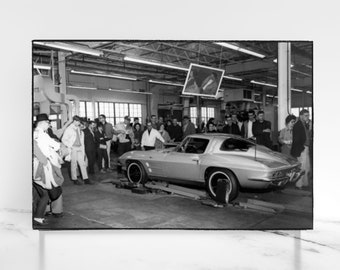 May include: A black and white photo of a group of people standing around a silver Corvette in a factory setting.