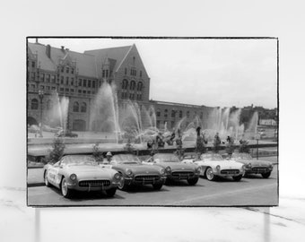 May include: Five classic black and white convertible sports cars parked in a row in front of a building with fountains. The cars are all facing the same direction and are lined up in a straight line.