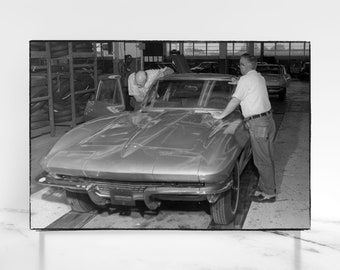 May include: Black and white photograph of a classic silver car in a factory setting. Two men are inspecting the car, one leaning on the hood. The car has a sleek, aerodynamic design, with a low profile and rounded features. Tires and other cars are visible in the background.