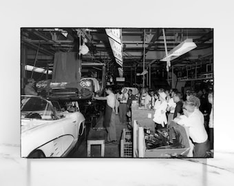 May include: Black and white photo of a car assembly line with workers watching a new car being lowered from the ceiling. The car is a classic Corvette.