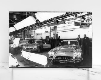 May include: Black and white photo of a car assembly line with two classic cars, one red and one light blue, in the foreground. The cars are being inspected by men in suits.