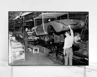 Op de afbeelding: Zwart-witfoto van een autoassemblagelijn. Werknemers zijn bezig met het assembleren van een rode auto. De auto ligt ondersteboven op een transportband. De foto is genomen vanuit een lage hoek.