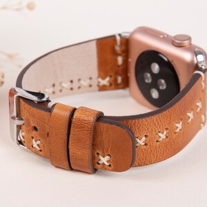 May include: A brown leather watch band with white stitching. The band is designed for an Apple Watch and has a silver buckle.