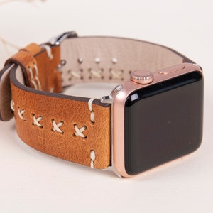 May include: A brown leather watch band with white stitching for an Apple Watch. The watch band has a rose gold Apple Watch attached to it.