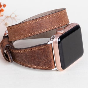 Puede incluir: Una correa de Apple Watch de cuero marrón con una esfera de reloj de oro rosa. La correa está envuelta dos veces y tiene un diseño cosido.