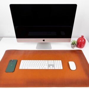Op de afbeelding: Een bruine leren bureau mat met een wit toetsenbord, een witte muis en een groene smartphone erop. De mat ligt op een wit bureau met een computermonitor en een rode vaas met een plant op de achtergrond.