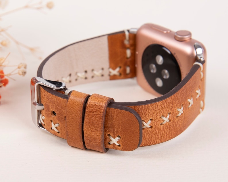 May include: A brown leather watch band with white stitching. The band is designed for an Apple Watch and has a silver buckle.