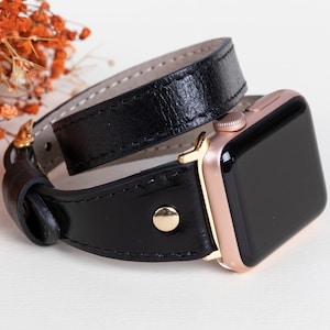 Puede incluir: Una correa de reloj de doble vuelta de cuero negro con detalles dorados, diseñada para un Apple Watch.