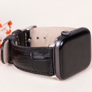 Puede incluir: Un reloj inteligente negro con una correa de cuero negro con textura de piel de cocodrilo. El reloj tiene una pantalla negra y un marco plateado. La correa se asegura con una hebilla plateada. El reloj se muestra sobre un fondo claro.