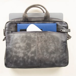 Op de afbeelding: Een grijze leren laptoptas met een ritsvak aan de voorkant. De tas is open en er is een laptop, een blauw notitieboekje en een wit voorwerp zichtbaar.