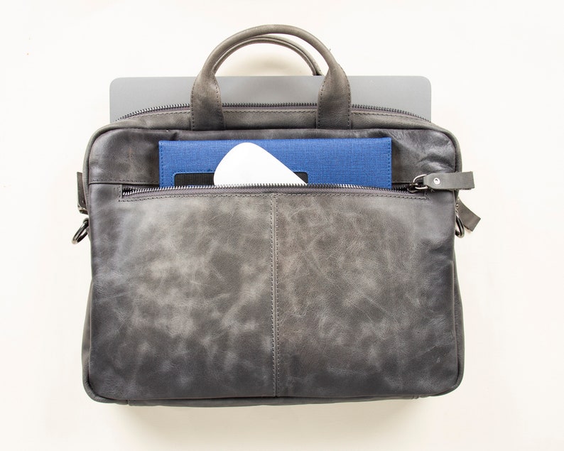 Op de afbeelding: Een grijze leren laptoptas met een ritsvak aan de voorkant. De tas is open en er is een laptop, een blauw notitieboekje en een wit voorwerp zichtbaar.