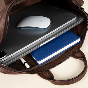 Op de afbeelding: Een bruine leren tas met een ritssluiting, open om een ​​laptop, een draadloze muis, een blauw notitieboekje en een witte stylus te laten zien.