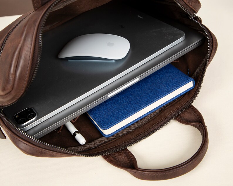 Op de afbeelding: Een bruine leren tas met een ritssluiting, open om een ​​laptop, een draadloze muis, een blauw notitieboekje en een witte stylus te laten zien.