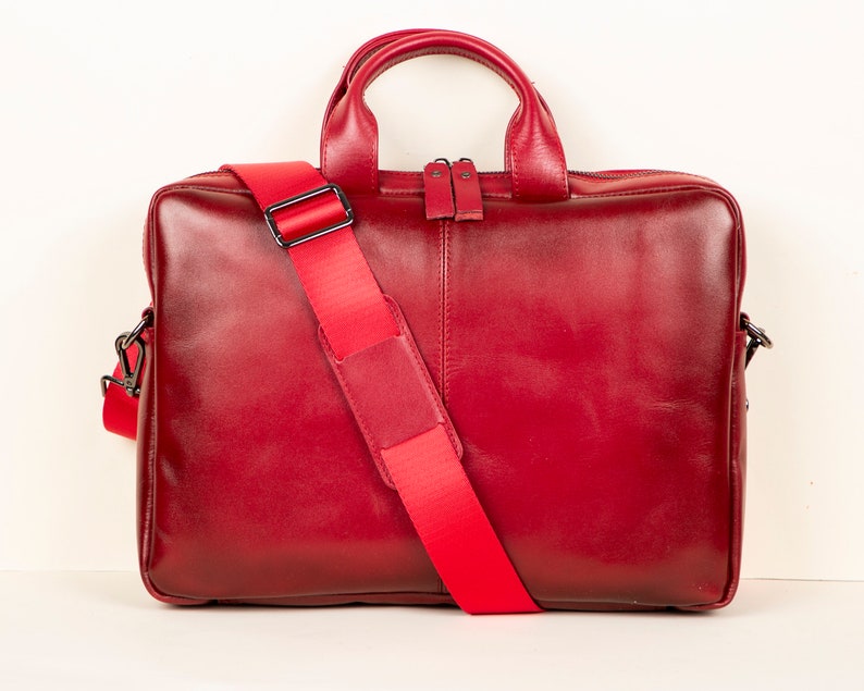 Op de afbeelding: Een rode leren laptoptas met een lange verstelbare schouderband. De tas heeft een bovenste handvat en een ritssluiting.