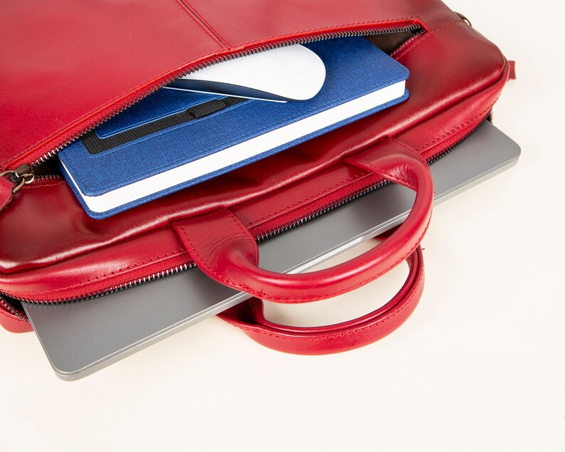 Op de afbeelding: Een rode leren laptoptas met een ritssluiting en een handvat. De tas is open en er zit een laptop, een notitieboekje en een draadloze muis in.