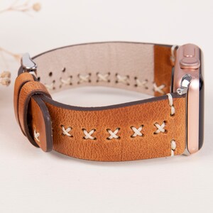 May include: A brown leather watch band with white stitching. The band has a stitched pattern of X's. The band is attached to a rose gold Apple Watch.